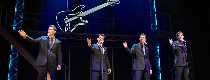 Four performers in matching dark suits stand at microphones onstage in a scene from Jersey Boys. Blue lighting and a large illuminated guitar outline frame the concert-style performance.