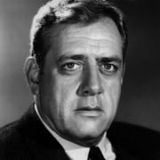 Black-and-white head-and-shoulders portrait of a person in a dark suit and tie, looking directly at the camera against a softly lit background.