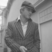 Black-and-white photograph of a person wearing a striped jacket, open-collar shirt, and flat cap, standing outdoors in an alleyway between buildings.