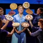 A performer in a light blue diner uniform stands center stage while four ensemble members surround the performer, each holding up a different pie. The pies frame the central figure against a backdrop of diner windows, creating a theatrical tableau from Waitress.