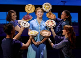 A performer in a light blue diner uniform stands center stage while four ensemble members surround the performer, each holding up a different pie. The pies frame the central figure against a backdrop of diner windows, creating a theatrical tableau from Waitress.