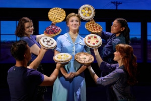 A performer in a light blue diner uniform stands center stage while four ensemble members surround the performer, each holding up a different pie. The pies frame the central figure against a backdrop of diner windows, creating a theatrical tableau from Waitress.