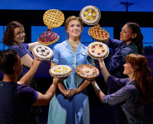 A performer in a light blue diner uniform stands center stage while four ensemble members surround the performer, each holding up a different pie. The pies frame the central figure against a backdrop of diner windows, creating a theatrical tableau from Waitress.