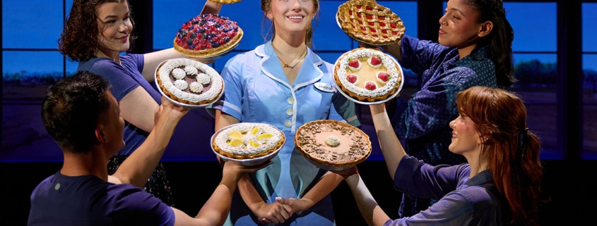 A performer in a light blue diner uniform stands center stage while four ensemble members surround the performer, each holding up a different pie. The pies frame the central figure against a backdrop of diner windows, creating a theatrical tableau from Waitress.
