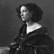 Black-and-white profile portrait of a young person with curly hair and earrings, wrapped in a dark draped garment and seated beside a table.