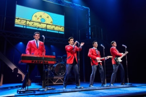 Four performers in matching red jackets perform at microphones during a concert scene from Jersey Boys. One stands at a keyboard while two play guitars, with a drummer and a colorful graphic of a record and musical notes displayed above the stage.