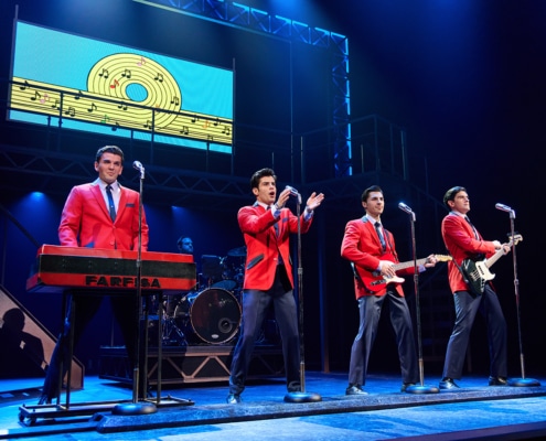 Four performers in matching red jackets perform at microphones during a concert scene from Jersey Boys. One stands at a keyboard while two play guitars, with a drummer and a colorful graphic of a record and musical notes displayed above the stage.