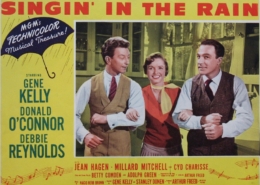Color lobby card for Singin’ in the Rain featuring three performers linked arm in arm in the center of the image. The design includes a yellow background, a blue umbrella graphic with the words “MGM’s Technicolor Musical Treasure!,” and prominent red title text reading “Singin’ in the Rain.” Cast and production credits appear along the sides and bottom of the promotional artwork.