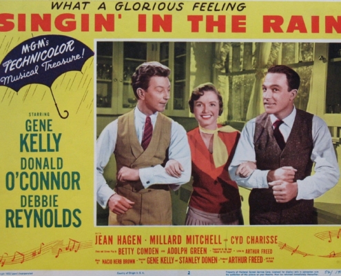 Color lobby card for Singin’ in the Rain featuring three performers linked arm in arm in the center of the image. The design includes a yellow background, a blue umbrella graphic with the words “MGM’s Technicolor Musical Treasure!,” and prominent red title text reading “Singin’ in the Rain.” Cast and production credits appear along the sides and bottom of the promotional artwork.