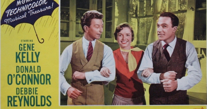Color lobby card for Singin’ in the Rain featuring three performers linked arm in arm in the center of the image. The design includes a yellow background, a blue umbrella graphic with the words “MGM’s Technicolor Musical Treasure!,” and prominent red title text reading “Singin’ in the Rain.” Cast and production credits appear along the sides and bottom of the promotional artwork.