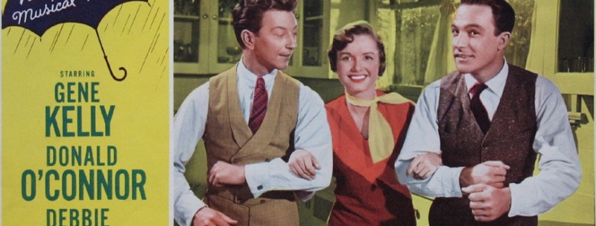 Color lobby card for Singin’ in the Rain featuring three performers linked arm in arm in the center of the image. The design includes a yellow background, a blue umbrella graphic with the words “MGM’s Technicolor Musical Treasure!,” and prominent red title text reading “Singin’ in the Rain.” Cast and production credits appear along the sides and bottom of the promotional artwork.