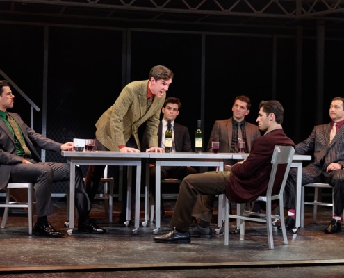 Six performers gather around two tables onstage in a tense meeting scene from Jersey Boys. One performer stands and leans forward over the table while the others remain seated, with wine bottles and glasses arranged between them.
