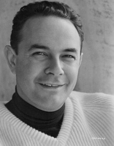 Black-and-white portrait of filmmaker Stanley Donen wearing a light-colored ribbed sweater over a dark turtleneck. Donen is shown from the shoulders up, looking slightly to the side against a softly textured background.
