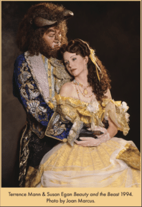 Studio publicity photo of two performers in elaborate stage costumes from Beauty and the Beast. One wears the Beast’s horned costume and embroidered blue coat, while the other wears Belle’s yellow ball gown, jewelry, and gloves as they pose together against a dark backdrop.