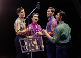 Four performers stand around a vintage studio microphone during a recording session scene from Jersey Boys. Wearing colorful patterned shirts, they sing together beside a music stand under warm stage lighting.