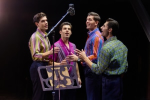 Four performers stand around a vintage studio microphone during a recording session scene from Jersey Boys. Wearing colorful patterned shirts, they sing together beside a music stand under warm stage lighting.
