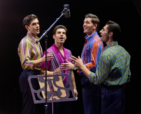 Four performers stand around a vintage studio microphone during a recording session scene from Jersey Boys. Wearing colorful patterned shirts, they sing together beside a music stand under warm stage lighting.