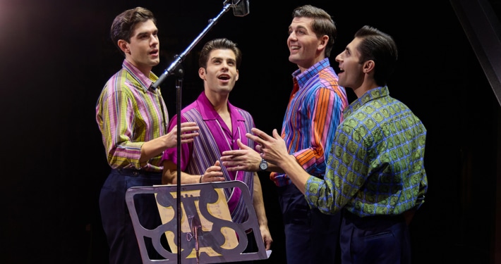 Four performers stand around a vintage studio microphone during a recording session scene from Jersey Boys. Wearing colorful patterned shirts, they sing together beside a music stand under warm stage lighting.