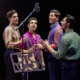 Four performers stand around a vintage studio microphone during a recording session scene from Jersey Boys. Wearing colorful patterned shirts, they sing together beside a music stand under warm stage lighting.
