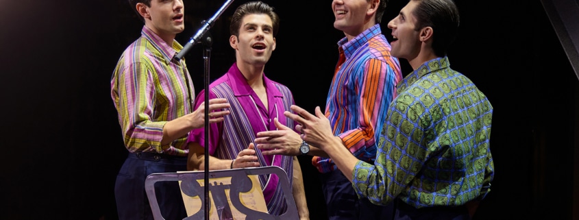 Four performers stand around a vintage studio microphone during a recording session scene from Jersey Boys. Wearing colorful patterned shirts, they sing together beside a music stand under warm stage lighting.