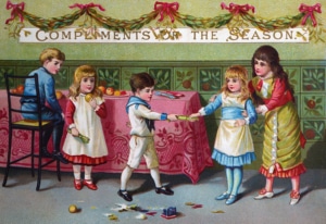 Victorian Christmas card showing four children gathered around a decorated table beneath a banner reading “Compliments of the Season.” Two children pull a Christmas cracker while another sits on a chair and a fourth stands nearby. Broken cracker contents and toys are scattered on the floor, with greenery garlands and red bows decorating the room.