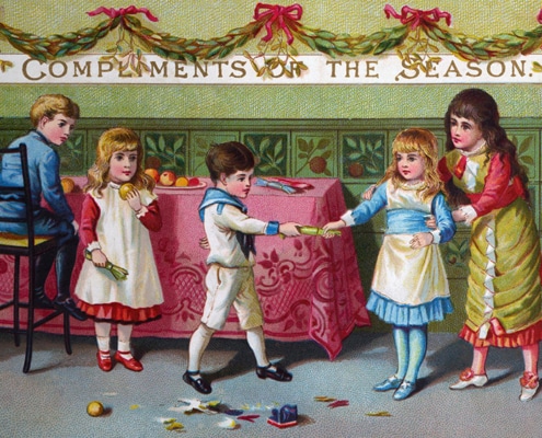Victorian Christmas card showing four children gathered around a decorated table beneath a banner reading “Compliments of the Season.” Two children pull a Christmas cracker while another sits on a chair and a fourth stands nearby. Broken cracker contents and toys are scattered on the floor, with greenery garlands and red bows decorating the room.