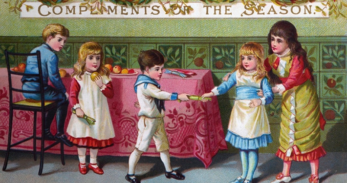 Victorian Christmas card showing four children gathered around a decorated table beneath a banner reading “Compliments of the Season.” Two children pull a Christmas cracker while another sits on a chair and a fourth stands nearby. Broken cracker contents and toys are scattered on the floor, with greenery garlands and red bows decorating the room.