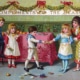 Victorian Christmas card showing four children gathered around a decorated table beneath a banner reading “Compliments of the Season.” Two children pull a Christmas cracker while another sits on a chair and a fourth stands nearby. Broken cracker contents and toys are scattered on the floor, with greenery garlands and red bows decorating the room.
