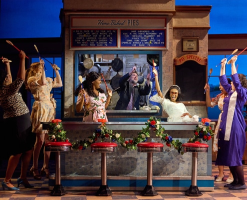 Ensemble members gather around the counter of a diner set in a scene from Waitress, raising wooden spoons overhead in a choreographed moment. A cook stands behind the service window beneath a menu board and “Pie Special” sign, with red diner stools lined across the counter.