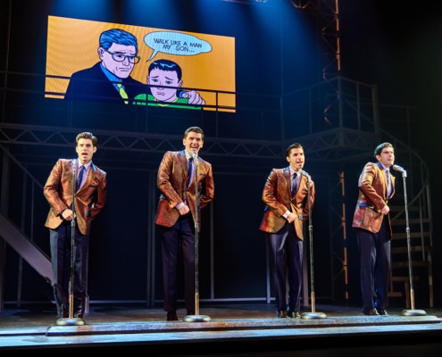 Four performers in matching bronze jackets sing and dance at standing microphones during a concert scene from Jersey Boys. Above them, a large comic-style graphic displays an illustration with the words “Walk Like a Man, My Son…”