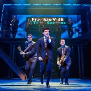 Three performers in dark suits perform at the front of the stage during a scene from Jersey Boys, with one singing into a handheld microphone and two playing electric guitars. A brass section performs on an elevated platform beneath a sign reading “Frankie Valli & The Four Seasons.”