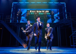 Three performers in dark suits perform at the front of the stage during a scene from Jersey Boys, with one singing into a handheld microphone and two playing electric guitars. A brass section performs on an elevated platform beneath a sign reading “Frankie Valli & The Four Seasons.”