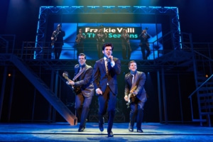 Three performers in dark suits perform at the front of the stage during a scene from Jersey Boys, with one singing into a handheld microphone and two playing electric guitars. A brass section performs on an elevated platform beneath a sign reading “Frankie Valli & The Four Seasons.”