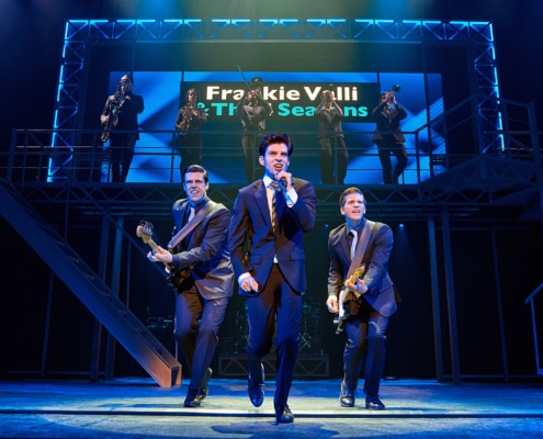 Three performers in dark suits perform at the front of the stage during a scene from Jersey Boys, with one singing into a handheld microphone and two playing electric guitars. A brass section performs on an elevated platform beneath a sign reading “Frankie Valli & The Four Seasons.”