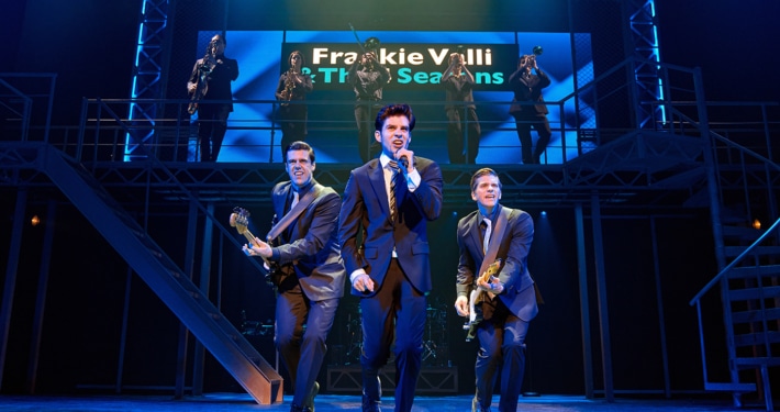 Three performers in dark suits perform at the front of the stage during a scene from Jersey Boys, with one singing into a handheld microphone and two playing electric guitars. A brass section performs on an elevated platform beneath a sign reading “Frankie Valli & The Four Seasons.”