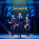 Three performers in dark suits perform at the front of the stage during a scene from Jersey Boys, with one singing into a handheld microphone and two playing electric guitars. A brass section performs on an elevated platform beneath a sign reading “Frankie Valli & The Four Seasons.”
