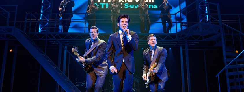 Three performers in dark suits perform at the front of the stage during a scene from Jersey Boys, with one singing into a handheld microphone and two playing electric guitars. A brass section performs on an elevated platform beneath a sign reading “Frankie Valli & The Four Seasons.”