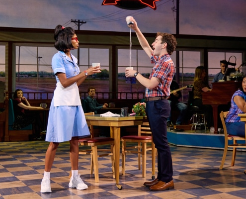 A scene from Waitress shows two performers in a diner. One, dressed in a light blue waitress uniform, holds a coffee mug while another performer in a plaid shirt playfully pours sugar from a container into a cup. Other diner patrons and musicians are visible in the background, with café tables, checkerboard flooring, and large windows framing a rural landscape.