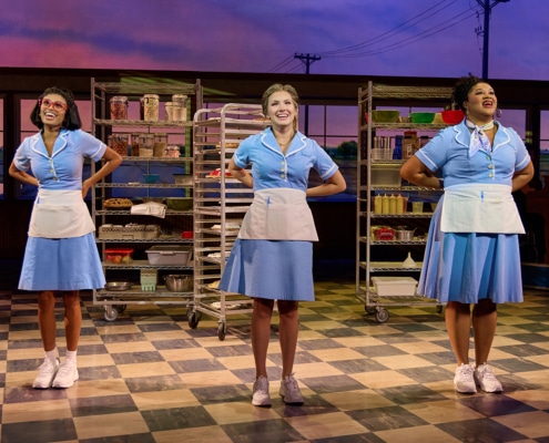 Three performers in matching light blue waitress uniforms stand in formation on a diner set during a scene from Waitress. Behind them are rolling bakery racks stocked with pies, baking supplies, and ingredients. The stage features a checkered floor, large windows, and warm theatrical lighting.