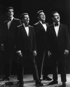 Black-and-white promotional photograph of four members of The Four Seasons standing together in tuxedos and bow ties onstage, posed beneath theatrical lighting.