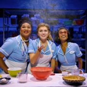 Three performers in light blue diner uniforms gather around a baking station in Waitress. Mixing bowls, measuring cups, ingredients, and a pie crust sit on the table as flour falls from above. Metal bakery racks filled with baking supplies are visible in the background.