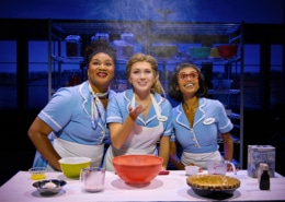Three performers in light blue diner uniforms gather around a baking station in Waitress. Mixing bowls, measuring cups, ingredients, and a pie crust sit on the table as flour falls from above. Metal bakery racks filled with baking supplies are visible in the background.