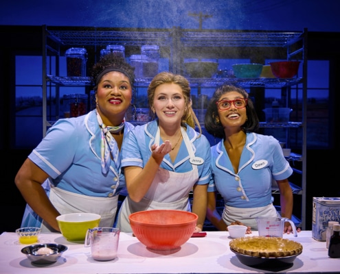 Three performers in light blue diner uniforms gather around a baking station in Waitress. Mixing bowls, measuring cups, ingredients, and a pie crust sit on the table as flour falls from above. Metal bakery racks filled with baking supplies are visible in the background.