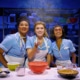 Three performers in light blue diner uniforms gather around a baking station in Waitress. Mixing bowls, measuring cups, ingredients, and a pie crust sit on the table as flour falls from above. Metal bakery racks filled with baking supplies are visible in the background.