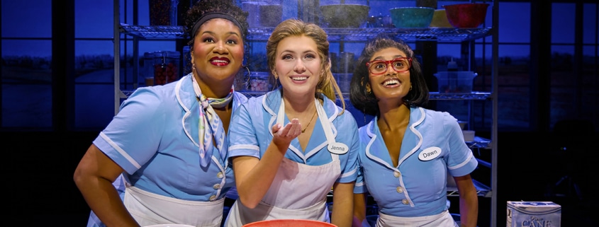 Three performers in light blue diner uniforms gather around a baking station in Waitress. Mixing bowls, measuring cups, ingredients, and a pie crust sit on the table as flour falls from above. Metal bakery racks filled with baking supplies are visible in the background.