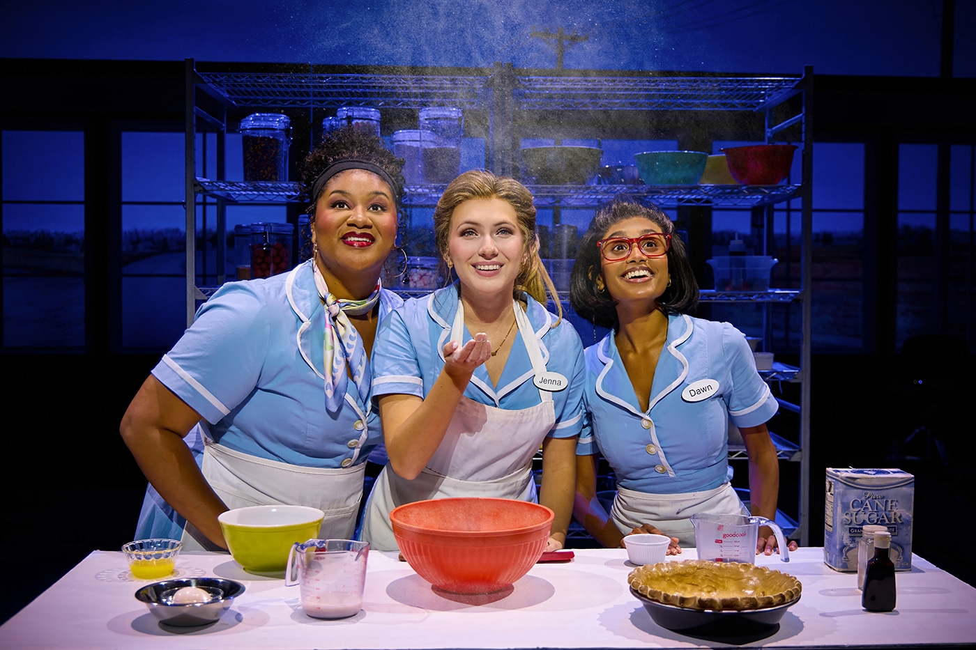 Three performers in light blue diner uniforms gather around a baking station in Waitress. Mixing bowls, measuring cups, ingredients, and a pie crust sit on the table as flour falls from above. Metal bakery racks filled with baking supplies are visible in the background.