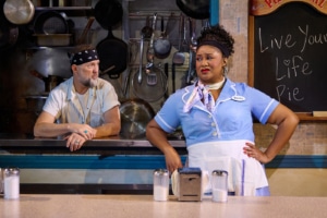 waitress in a light blue diner uniform stands at a restaurant counter with hands on hips while a cook leans on the service window behind the counter. The diner kitchen is filled with hanging cookware, and a chalkboard in the background reads “Live Your Life Pie.”