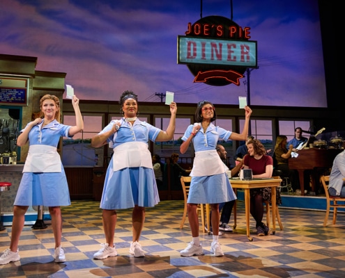 Three waitresses in matching light blue uniforms stand center stage holding order pads beneath a glowing “Joe’s Pie Diner” sign in a scene from Waitress. Diners sit at tables around them, and musicians perform on a raised platform in the background.