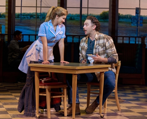 A waitress in a light blue diner uniform leans across a table toward a seated customer in a plaid shirt during a scene from Waitress. The customer holds a plate while the two converse in a diner setting with checkered flooring, booth seating, and large windows overlooking a rural backdrop.