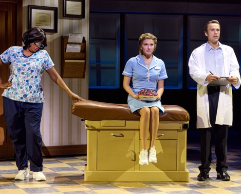 A performer in a light blue waitress uniform sits on an examination table holding a berry pie while flanked by two medical professionals, one in scrubs and one in a white lab coat. The scene takes place on a doctor's office set with framed certificates, an exam table, and large windows in the background. Provide your feedback on BizChat